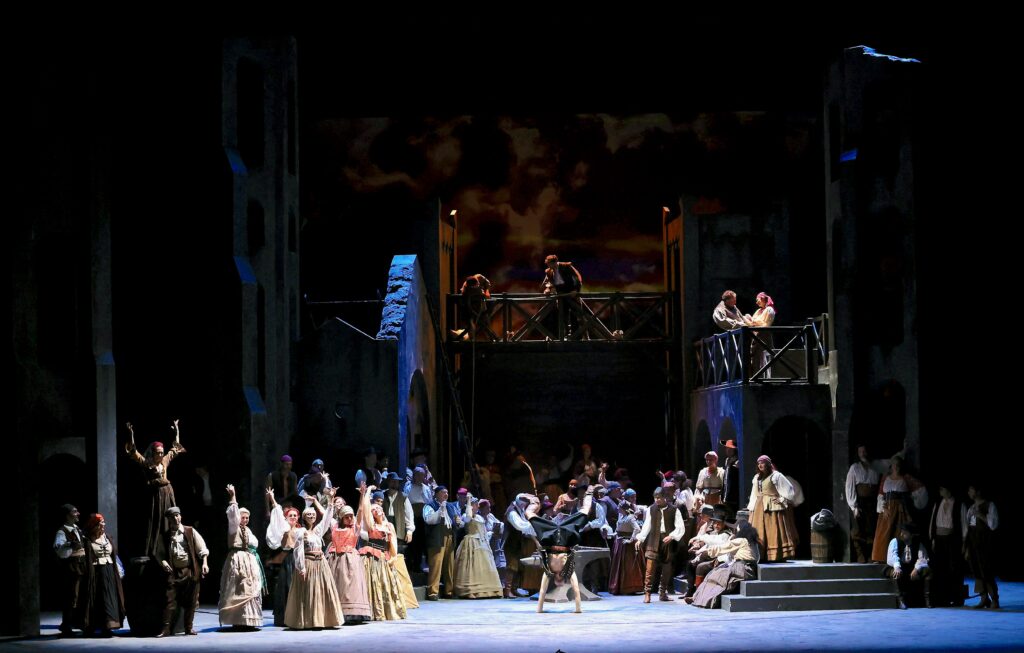 Foto d’insieme di una scena del Trovatore, con numerosi personaggi in costume disposti su più livelli scenografici e illuminati da luci